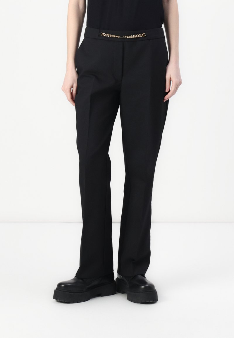 Zwarte broek met een getailleerde pasvorm, voorzien van een gouden kettingaccent bij de taille en een gladde textuur, gecombineerd met dikke zwarte schoenen.