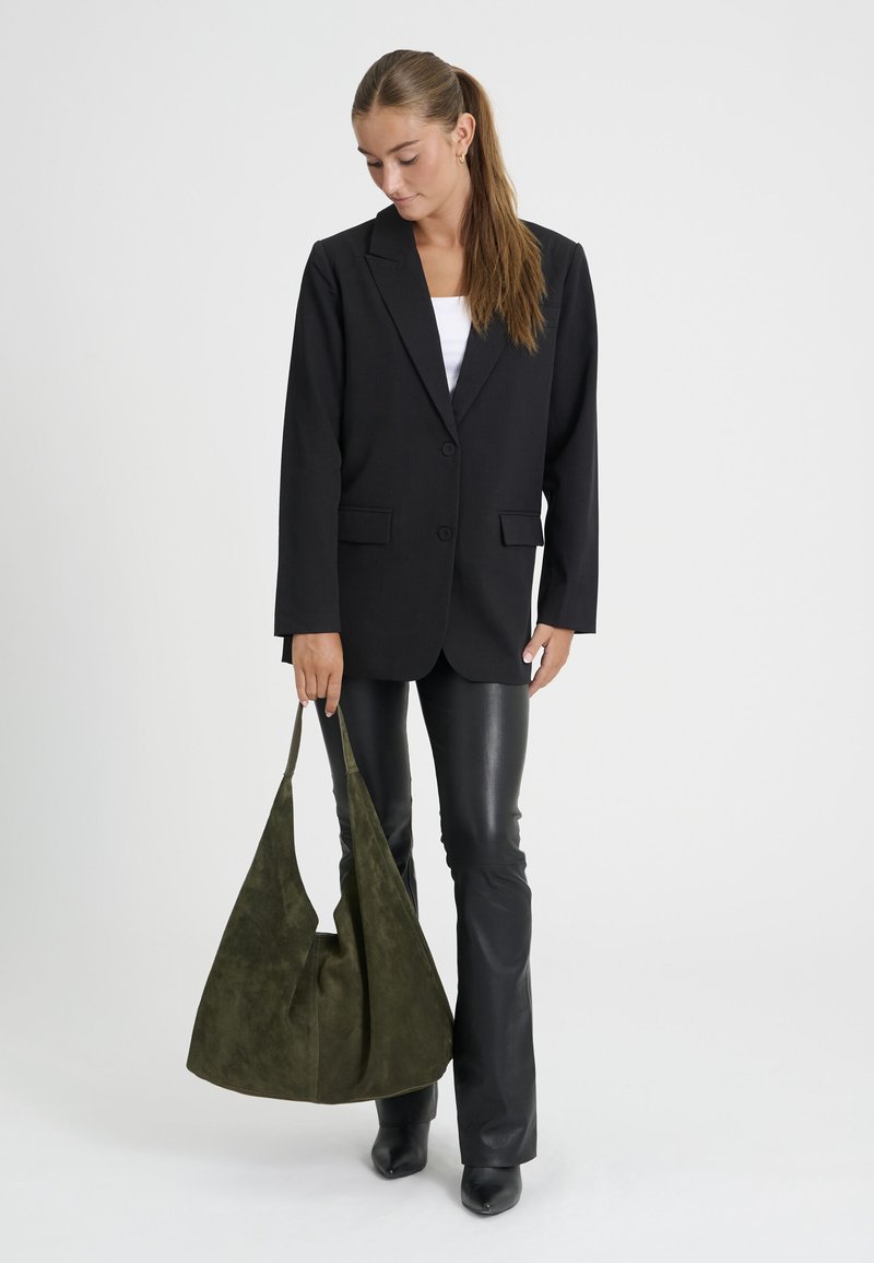 Femme portant un blazer noir et un pantalon en cuir tenant un grand sac à bandoulière en daim vert olive sur un fond uni.