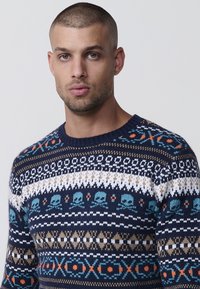 Temno modra pletena jopica s Fair Isle vzorom, ki vključuje lobanje, geometrijske oblike ter akcentne barve v turkizni, beli, oranžni in rjavi.
