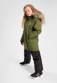 Oliwkowo-zielona parka z kapturem wyłożonym futerkiem, zapinana z przodu na zamek błyskawiczny, z bocznymi kieszeniami. Noszona na czarnych spodniach i uzupełniona czarnymi butami z wzorem w jodełkę.