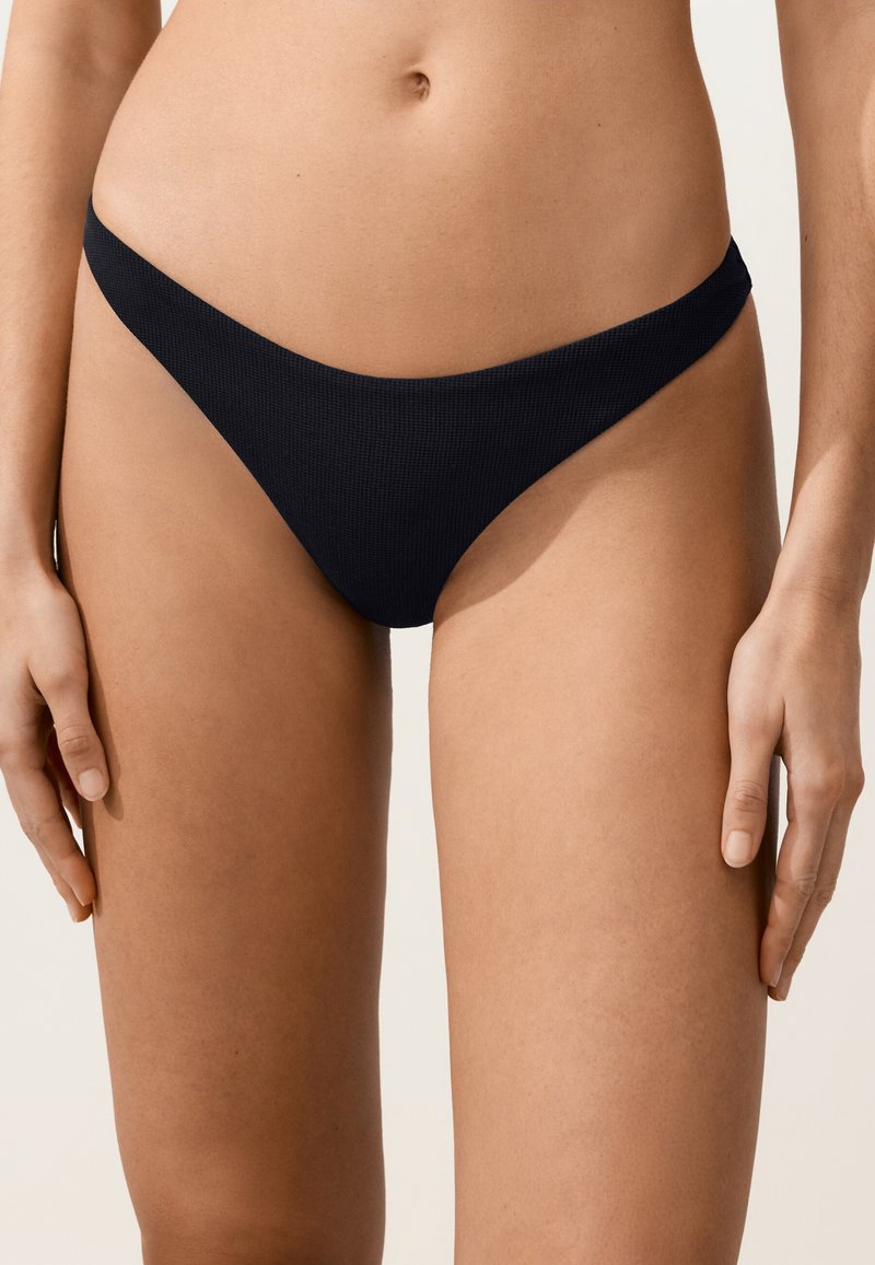 Nő fekete, texturált bikini alsórésszel, deréktól a comb közepéig látható, világos háttér előtt.