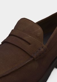 Καφέ μοκασίνι από σουέτ με λουράκι για κέρματα και εμφανή ραφές κατά μήκος του δακτύλου και της πλευράς, με μαύρη σόλα.