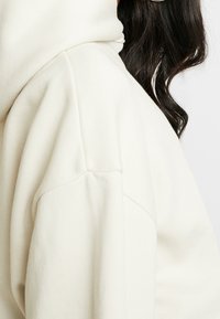 Kremowa bluza z kapturem z wysokim kołnierzem i opuszczonymi ramionami. Materiał ma miękką fakturę z widocznymi detalami przeszyć.