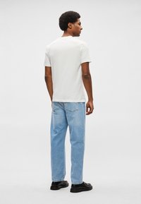 Balts īsās piedurknes T-krekls kopā ar gaiši ziliem taisnā griezuma džinsiem un melnām kurpēm, redzams no mugurpuses pret gaišu fonu.