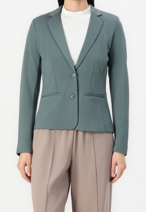Kvinde iført en tætsiddende salviegrøn blazer med to knapper over en hvid top og beige, plisserede bukser mod en hvid baggrund.