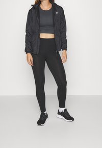 Zwarte lichte hooded jacket, nauwsluitend zwart sporttopje en hoge zwarte leggings. Een paar zwarte sneakers met witte zolen en sokken.