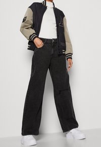 Svarta jeans med vida ben, matchade med en svart och beige varsityjacka och vita sneakers. Slät tyg med kontrasterande ärmar och emblem.