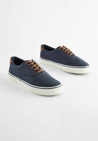 Marineblauwe canvas sneakers met bruine leren accentstukken, witte rubberen zool en contrasterende blauwe streep. Vetersluiting en een gestructureerd oppervlak.