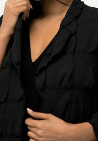 Black textured jacket with ruffled edges, featuring a V-neck design. The fabric shows a crinkled pattern and soft, lightweight appearance.