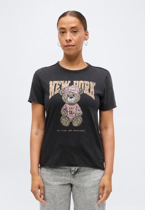 Γυναίκα με μαύρο μπλουζάκι που έχει αρκουδάκι με ροζ καπέλο και πουκάμισο, το κείμενο "NEW YORK" από πάνω και ανοιχτό γκρι τζιν, με φόντο μια απλή επιφάνεια.