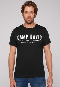 Camiseta de algodón negra con mangas cortas, con un texto blanco y audaz "CAMP DAVID" en el pecho y texto adicional abajo en fuente más pequeña.
