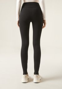 Leggings neri con un alto punto vita, texture liscia e design aderente, abbinati a scarpe da ginnastica chiare.