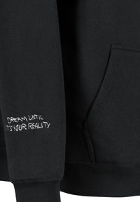 Schwarze Kapuzenjacke mit weicher Textur. Verfügt über eine Fronttasche und einen weißen gestickten Spruch auf dem Ärmel: "TRÄUME, BIS ES DEINE REALITÄT WIRD."