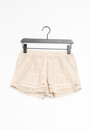 Short en dentelle beige au crochet avec taille élastique suspendu à un cintre en plastique noir sur fond blanc.
