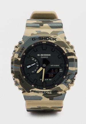 Casio G-Shock-klokke med kamuflasjemønster, analog-digital skjerm, dagsindikator og funksjonsknapper på beige bakgrunn.