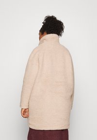 Beige fleecejacka med hög krage, med en texturerad yta och lös passform. Jackan går ner till mitten av låret.