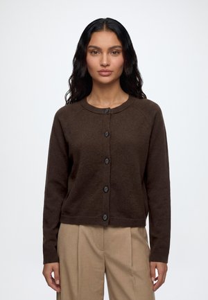 Donna con lunghi capelli scuri che indossa un cardigan marrone scuro abbottonato e pantaloni beige, in piedi davanti a uno sfondo chiaro e uniforme.