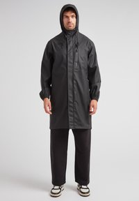 Schwarze wasserdichte Regenjacke mit Kapuze, Reißverschluss-Vordertasche, elastischen Bündchen und geradem Schnitt. Kombiniert mit dunklen Hosen und Sneakers.
