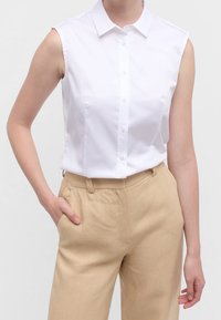 Chemise blanche sans manches avec col pointu, fermeture à boutons sur le devant et texture lisse, associée à un pantalon taille haute beige clair.