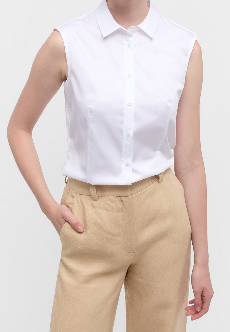 Chemise blanche sans manches avec col pointu, fermeture à boutons sur le devant et texture lisse, associée à un pantalon taille haute beige clair.