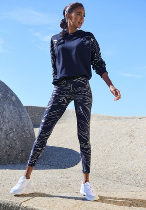 Navy hoodie with patterned sleeves, paired with form-fitting leggings featuring a swirling design in beige, and white athletic shoes.