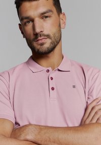 Poloshirt in lichtroze met een textuurstof, drie bordeauxrode knopen en een klein zwart logo op de linker bovenkant.