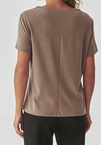 Camicia marrone a maniche corte con una texture morbida, scollatura rotonda e una cucitura centrale lungo la schiena, abbinata a pantaloni neri.