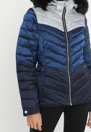 Frau in einer blauen, schwarzen und hellgrauen Daunenjacke mit einem Frontreißverschluss und einer Kapuze mit schwarzem Kunstfellrand.