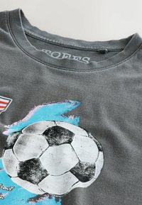 T-shirt gris avec un graphisme usé de ballon de football noir et blanc et un motif d'éclaboussures de peinture bleue près du col.