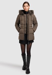 Vrouw in een bruine gewatteerde hooded jacket, zwarte mini skirt, sheer zwarte maillot en zwarte kniehoge laarzen, staand voor een effen achtergrond.