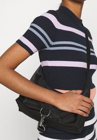 Musta satiinista valmistettu olkalaukku, jossa on kaareva muoto, metalliset yksityiskohdat ja säädettävä hihna, pidettynä kädessä raidallista toppia vasten.