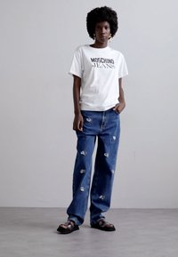 Balti marškinėliai su "Moschino Jeans" tekstu, derinami su laisvais mėlynais džinsais, turinčiais aiškių akcentų, ir juodomis šlepetėmis. Neutralus fonas.