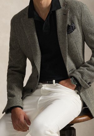 Homme portant un blazer en chevrons, un polo noir, un pantalon blanc et une montre-bracelet, assis sur un tabouret en bois avec une main dans la poche.