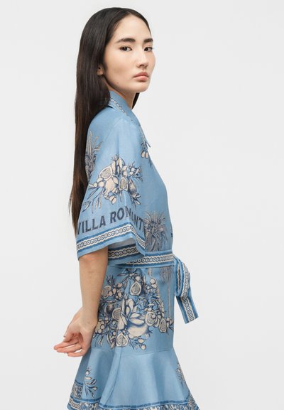 Νέα γυναίκα με μακριά σκουρόχρωμα μαλλιά που φοράει μπλε φόρεμα με μοτίβο λουλουδιών και κείμενο «Villa Romana», στέκεται πλαγίως με τα χέρια πίσω από την πλάτη.