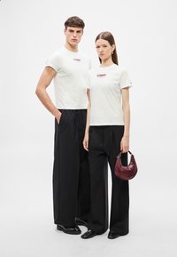Λευκά t-shirts με το λογότυπο "TOMMY", σε συνδυασμό με μαύρο παντελόνι φαρδύ σε ίσια γραμμή. Η γυναίκα κρατά μια μικρή μπορντό τσάντα χειρός με υφή.