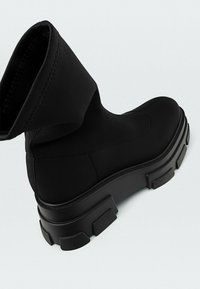 Czarne botki za kostkę z elastycznego materiału z grubą gumową podeszwą, charakteryzujące się eleganckim designem i subtelnymi detalami szycia.