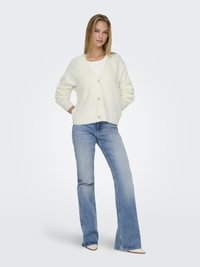 White knitted cardigan with a V-neck, five buttons, and puffed sleeves, paired with flared blue jeans and beige heeled shoes.