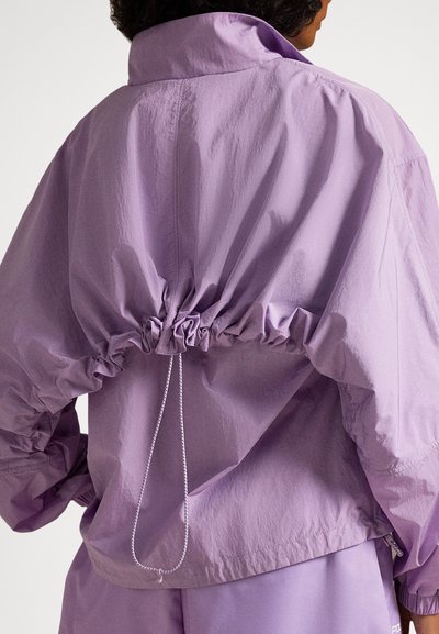 Πίσω όψη ατόμου που φοράει χαλαρό, ανοιχτό μοβ μπουφάν με ρυθμιζόμενο κορδόνι στο πάνω μέρος της πλάτης και αντίστοιχο παντελόνι.