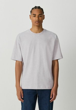 Junger Mann mit geflochtenem Haar trägt ein schlichtes hellgraues oversized T-Shirt und dunkelblaue Jeans vor schlichtem Hintergrund.
