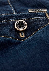 Donkerblauwe denimstof met gedetailleerde stiksels, een metalen knoop gegraveerd met "MEYER," en een klein rechthoekig metalen label dat ook "MEYER" is gebrandmerkt.