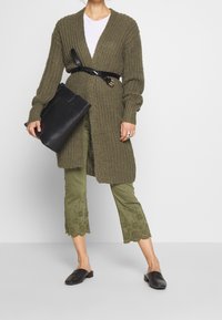 Knit olive green cardigan with a deep v-neck, black belt, black tote bag, green cropped pants with embroidered hem, and black slip-on shoes.