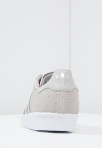 Sneaker grigio chiaro con tomaia in suede testurizzato, suola in gomma bianca e marchio metallico argento sul tallone. Le righe laterali presentano perforazioni.