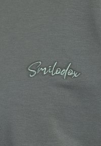 Camicia in tessuto a righe grigie con un logo ricamato blu chiaro "Smiledex" in posizione prominente. Texture liscia e design aderente.