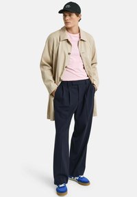 Beige trenssi takki vaaleanpunaisen t-paidan päällä, tummansiniset pleatatut housut ja siniset lenkkarit valkoisilla yksityiskohdilla. Musta lippalakki, jossa logo edessä.