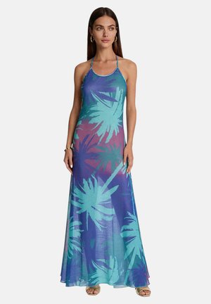 Donna che indossa un vestito senza maniche, lungo fino alla caviglia, con motivi di foglie tropicali blu e viola, in piedi con le braccia rilassate ai lati.