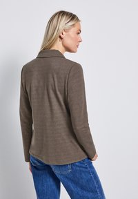 Chemise longue à manches brunes avec un col, confectionnée en tissu doux. Dotée d'un ourlet arrondi et d'une coupe décontractée, portée avec un jean bleu.