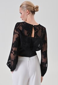 Zwarte doorschijnende blouse met een bloemenkantpatroon, lange klokmouwen en een sleutelgatopening aan de achterkant. Gecombineerd met witte broeken.