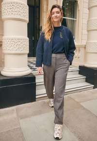 Navy textured bomber jacket over a blue sweatshirt, paired with light checkered trousers and beige sneakers, walking on stone steps.