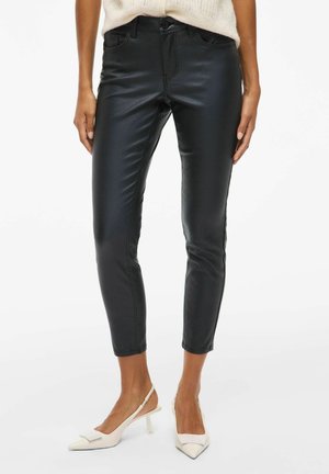 Zwarte namaak leren high-waisted broek met een strakke, glanzende textuur, voorzien van zijzakken en een slim-fit ontwerp, gecombineerd met witte hakken.
