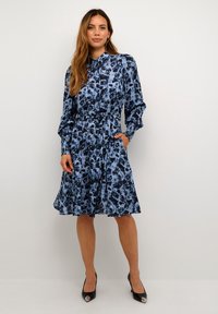 Rochie florală albastră cu guler, mâneci lungi, talie strânsă și buzunare, realizată dintr-un material ușor cu fustă fluidă.